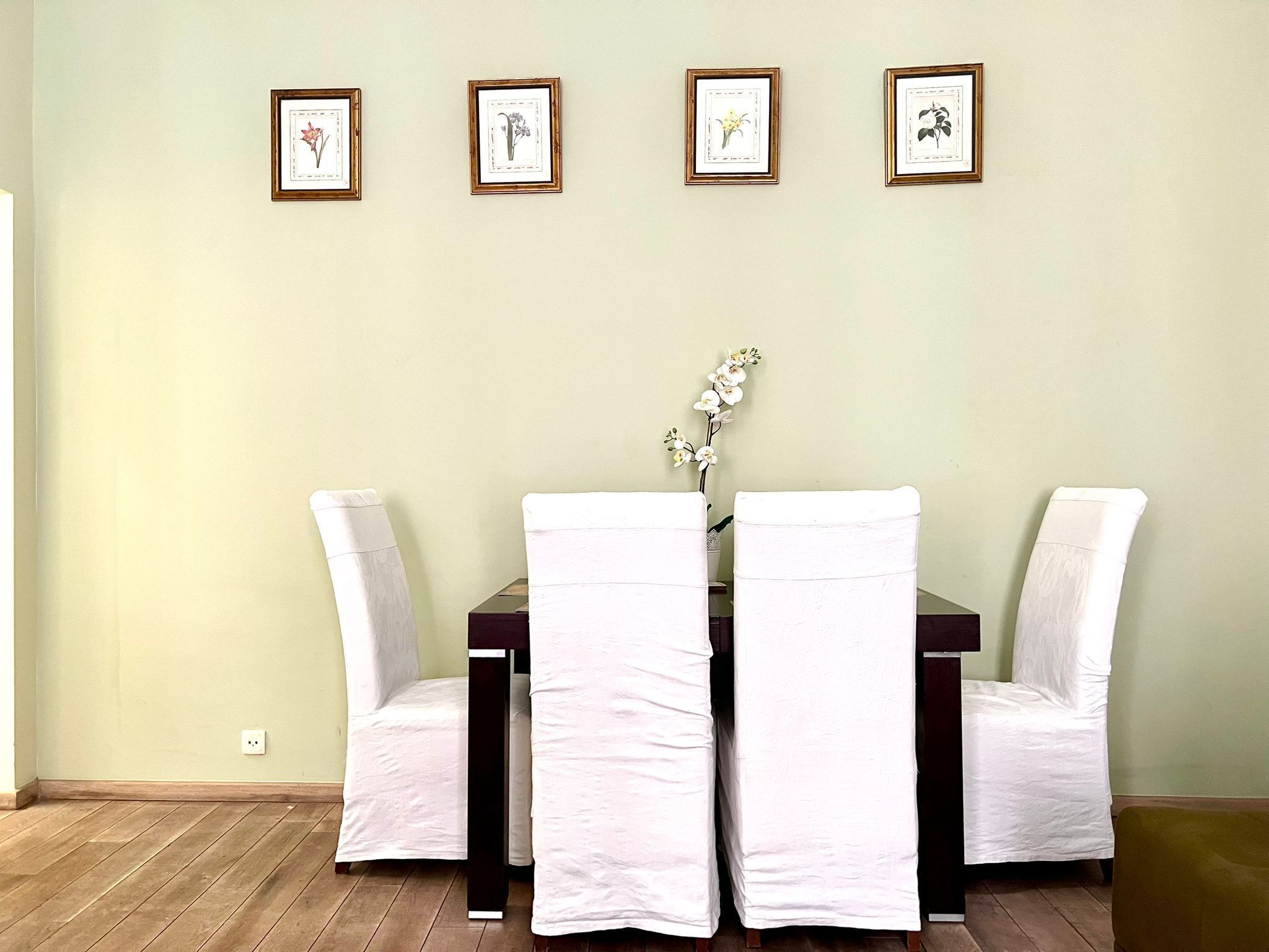 Een eethoek met een tafel en vier stoelen, elk met een witte hoes. Boven de tafel hangen vier ingelijste foto's.