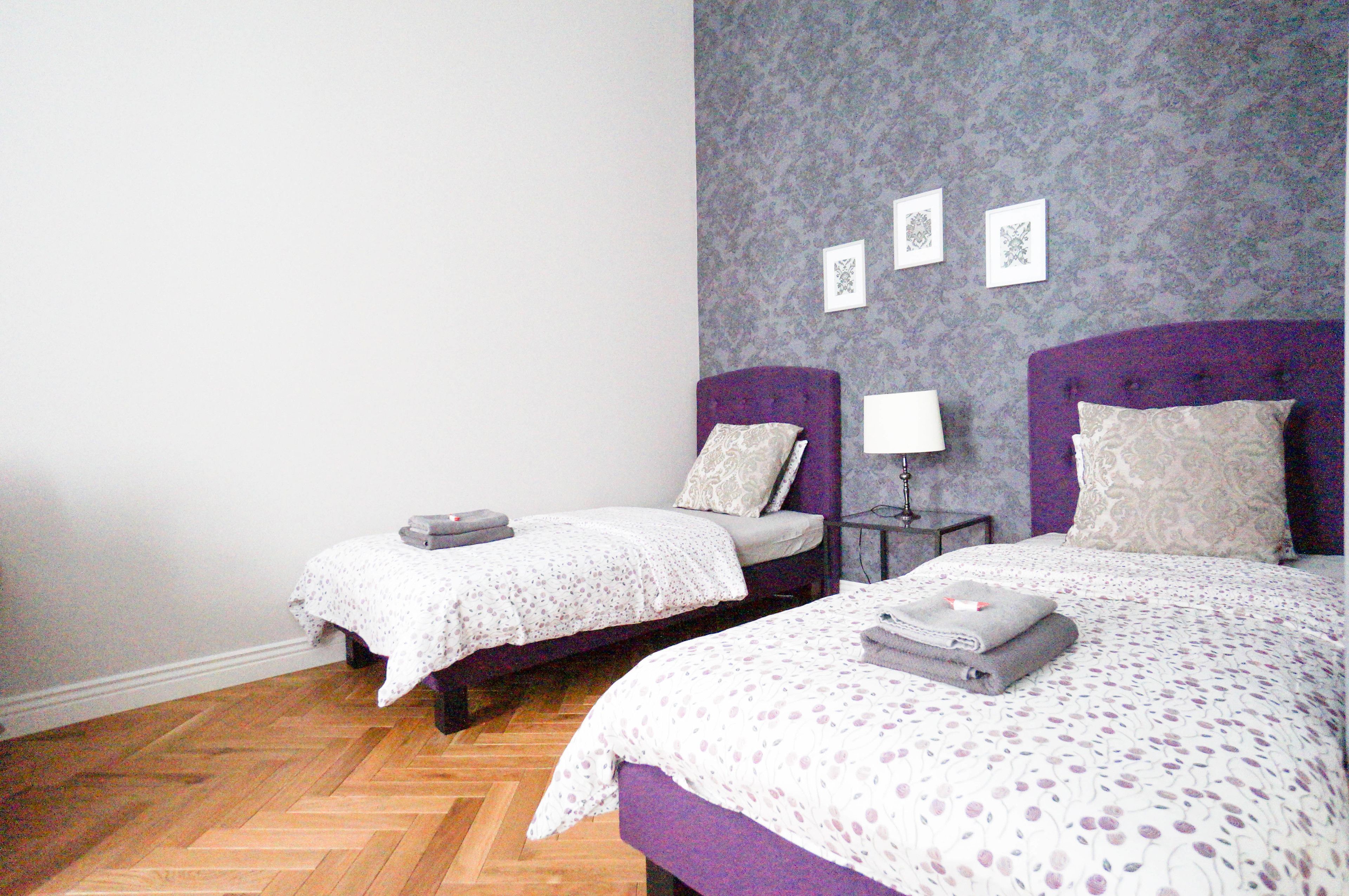 A room with two beds, each with a purple headboard, a small bedside table with a lamp, and a patterned wall.
