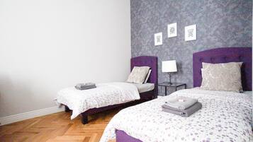 A room with two beds, each with a purple headboard, a small bedside table with a lamp, and a patterned wall.