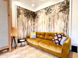 A room with a mustard-colored sofa, a small round table, a lamp, and a large abstract wall mural.