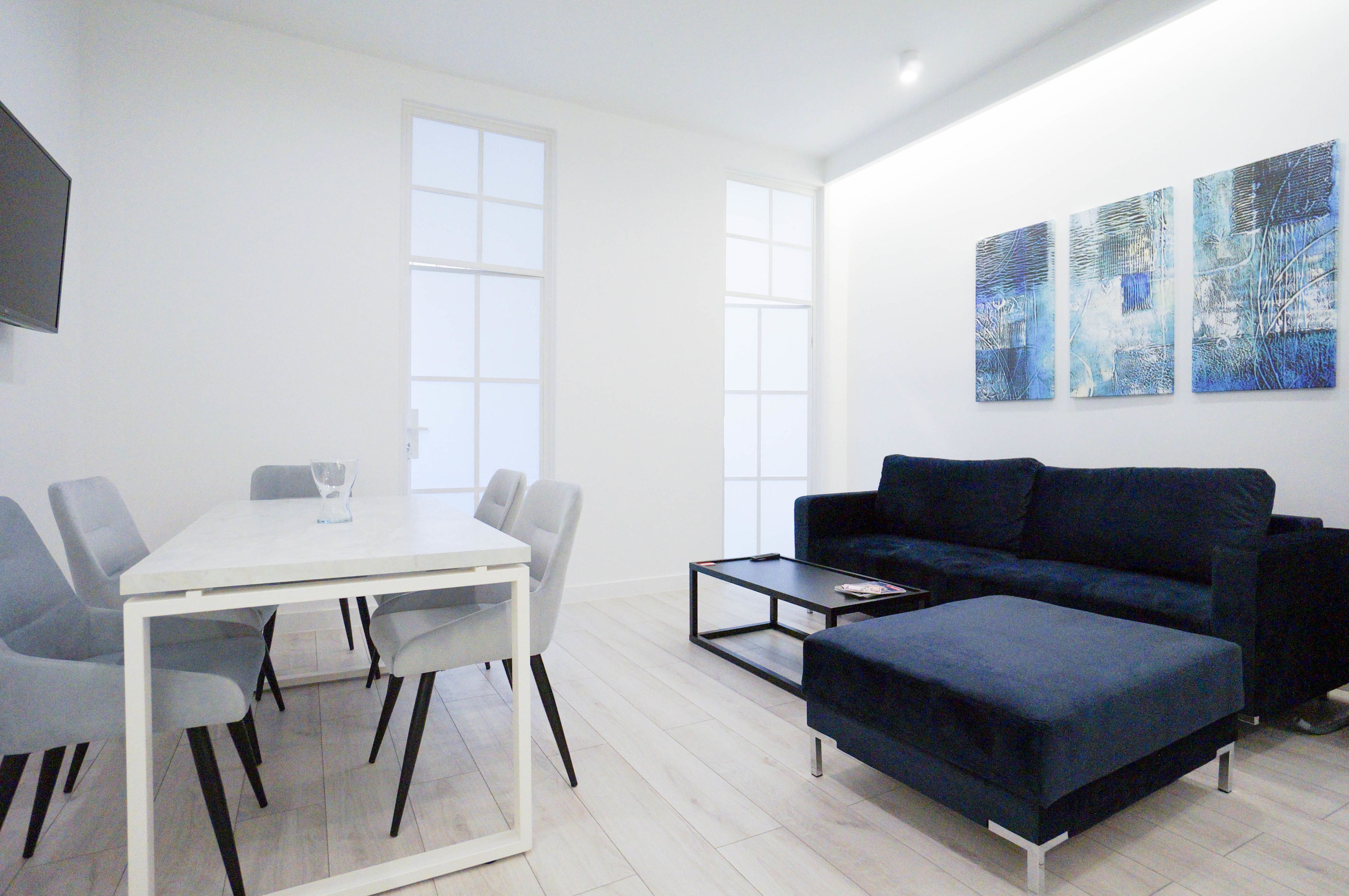Un salón moderno con una mesa de comedor blanca, sillas grises, un sofá azul oscuro y un otoman a juego. Hay cuadros abstractos en la pared y un televisor de pantalla plana montado allí.