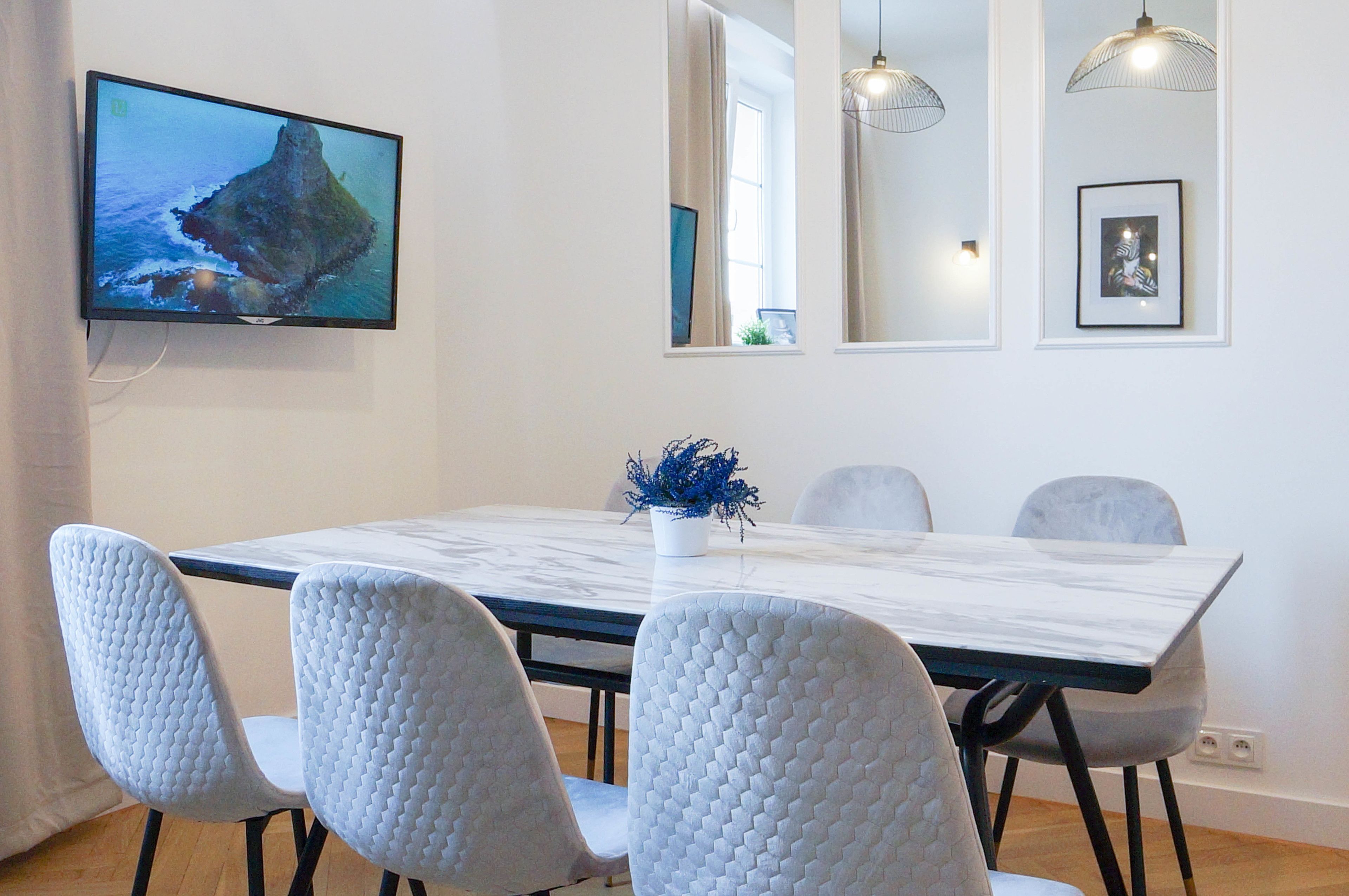 Un comedor con una mesa de mármol, cuatro sillas grises, un televisor en la pared y una planta decorativa.