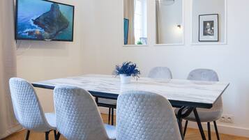 Un comedor con una mesa de mármol, cuatro sillas grises, un televisor en la pared y una planta decorativa.