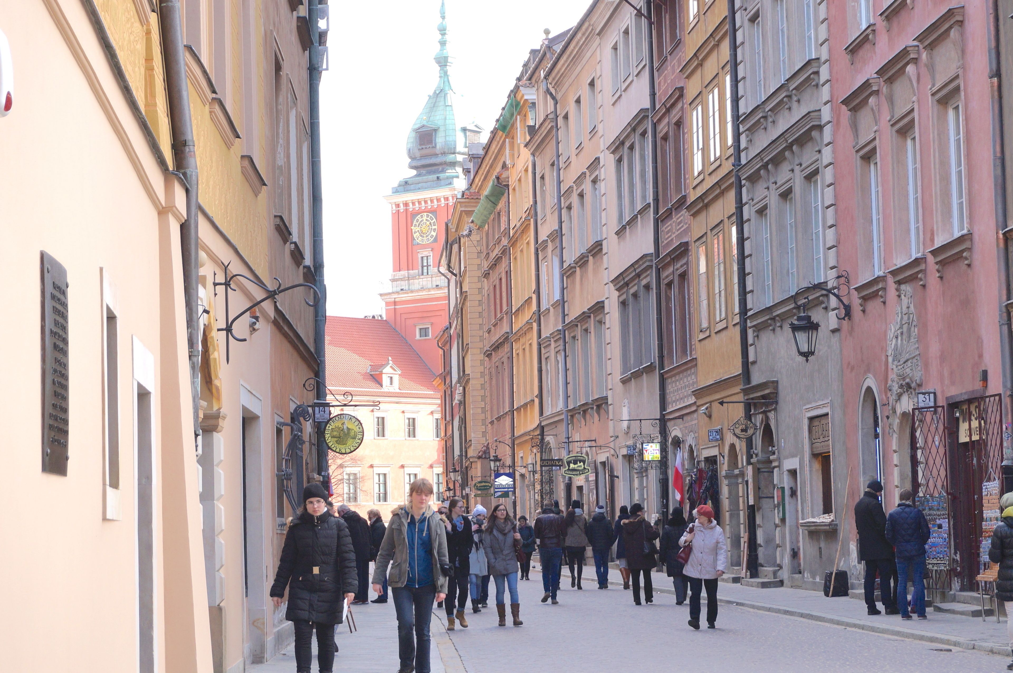 A narrow street lined with historic buildings, a prominent clock tower, and pedestrians.