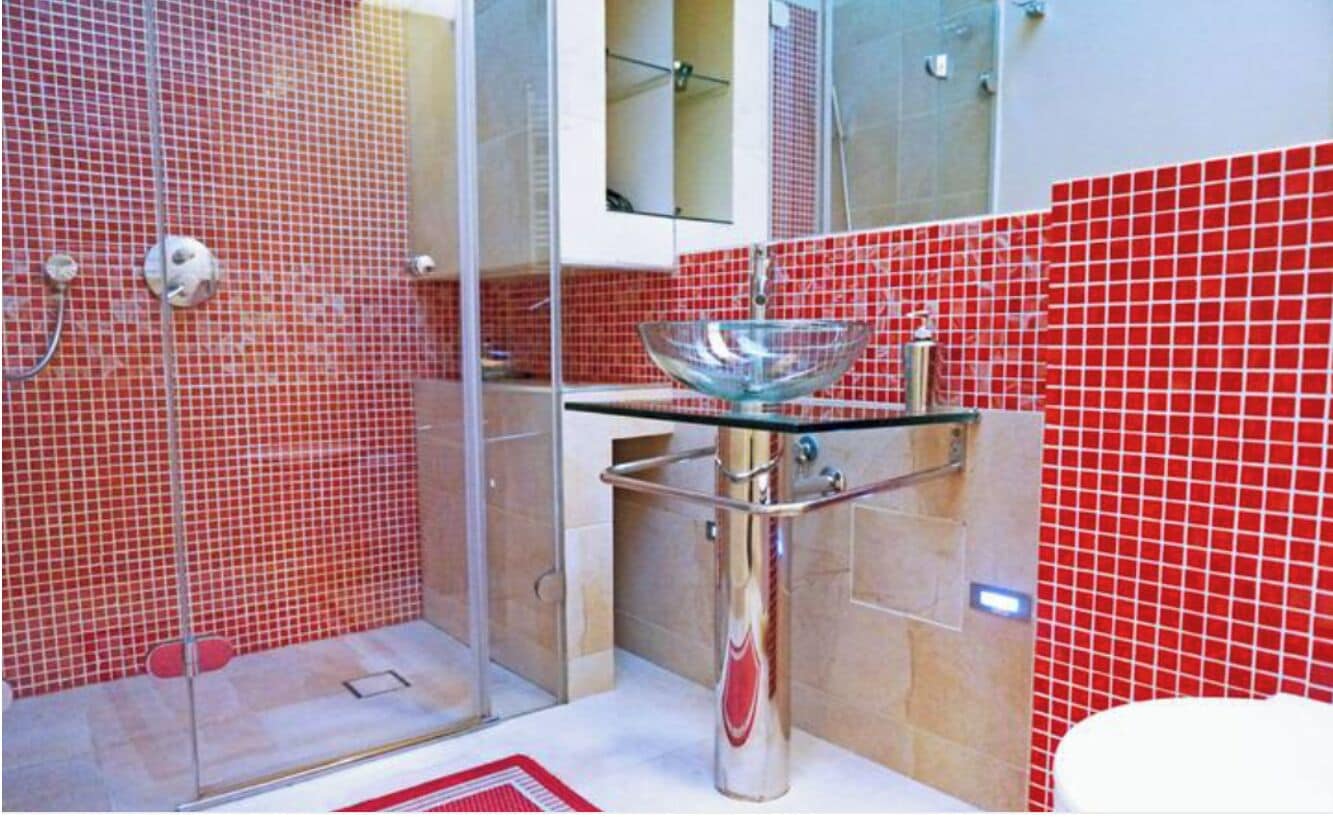 A bathroom with a glass shower enclosure, a glass sink, and red-tiled walls.