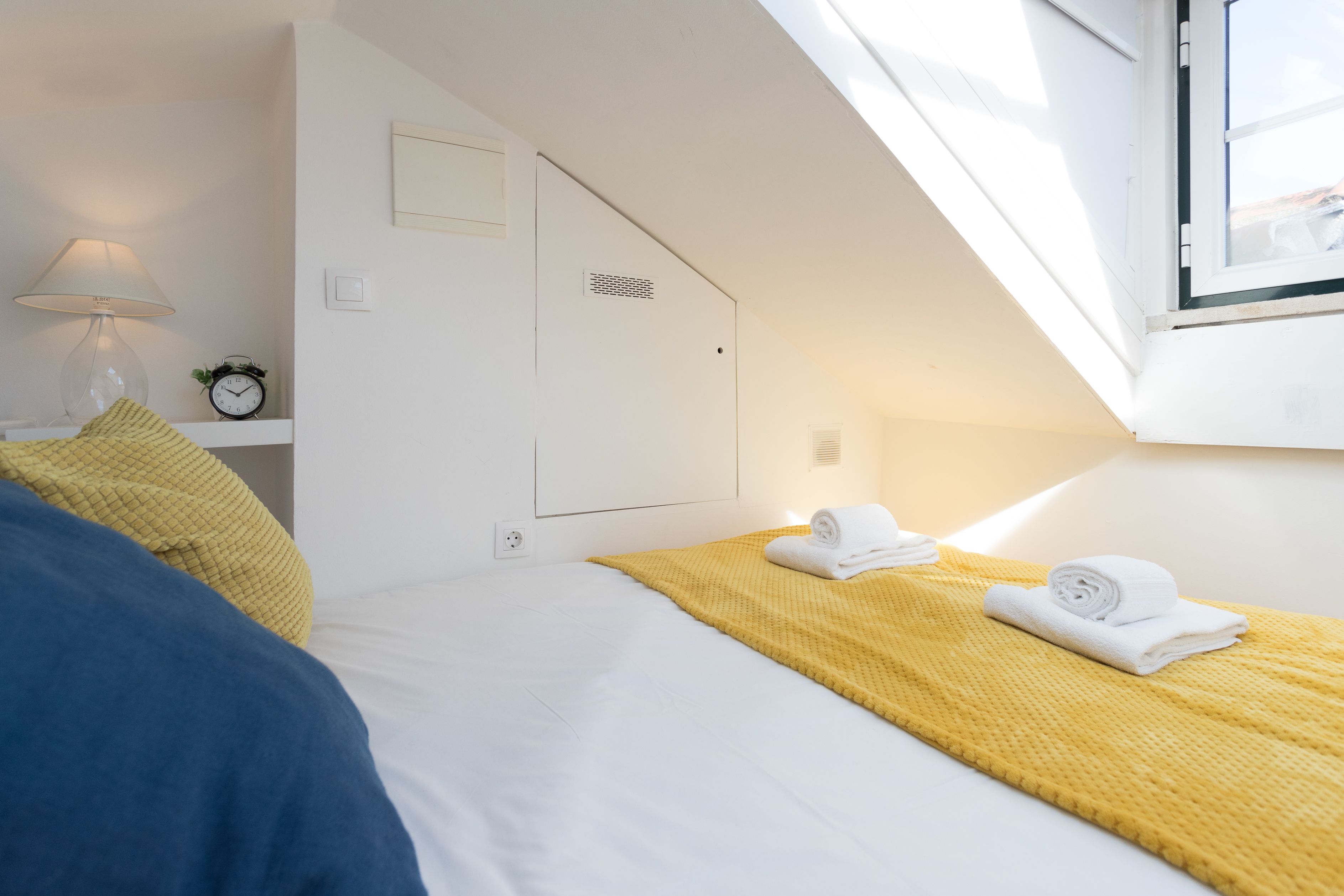 A bedroom with a bed, a yellow blanket, a white pillow, a lamp, and a clock on a bedside table.