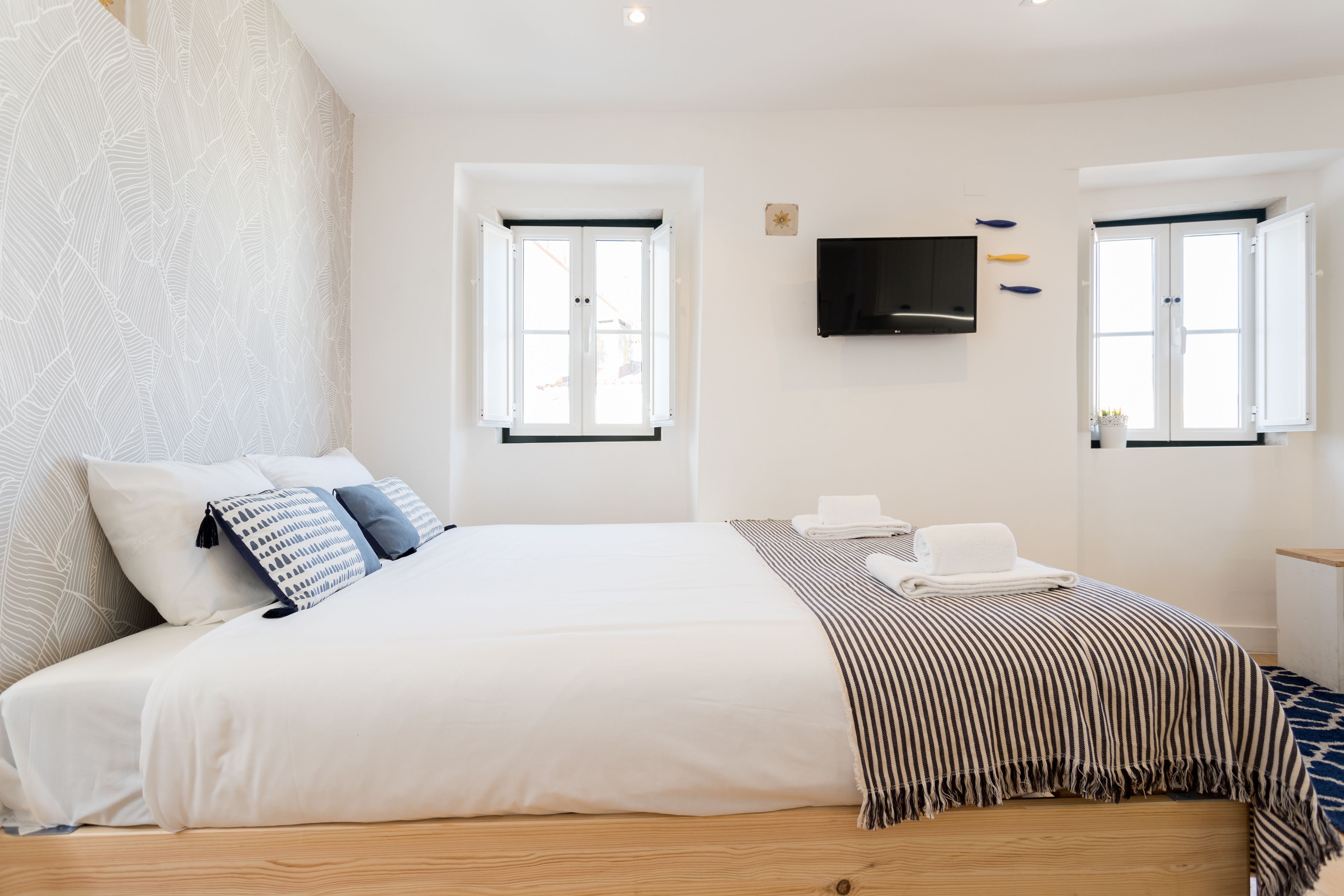 A modern hotel room with a large bed, two windows, a flat-screen TV, and a small table.