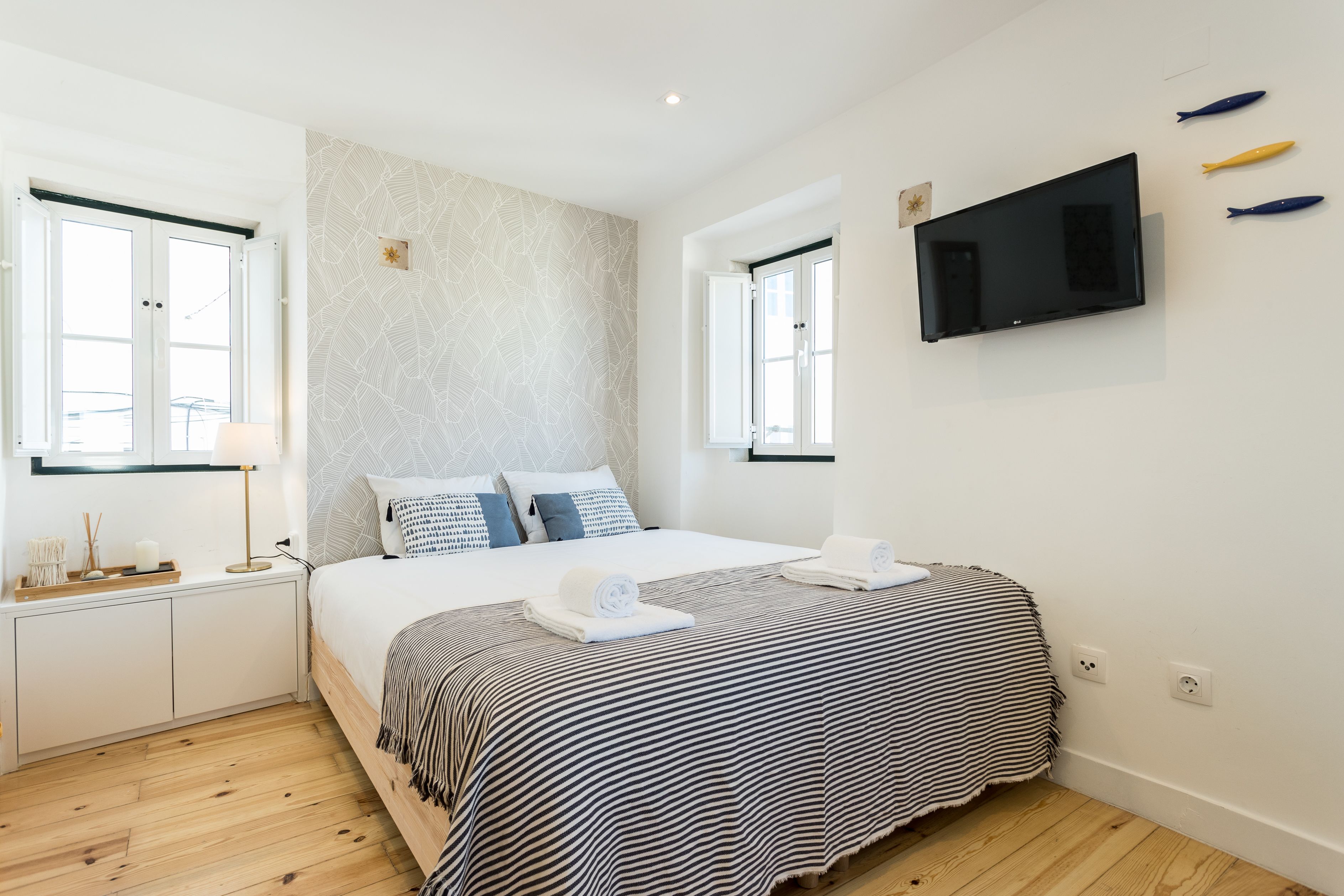 Quarto moderno com uma cama grande, duas janelas, uma televisão de tela plana e piso de madeira.