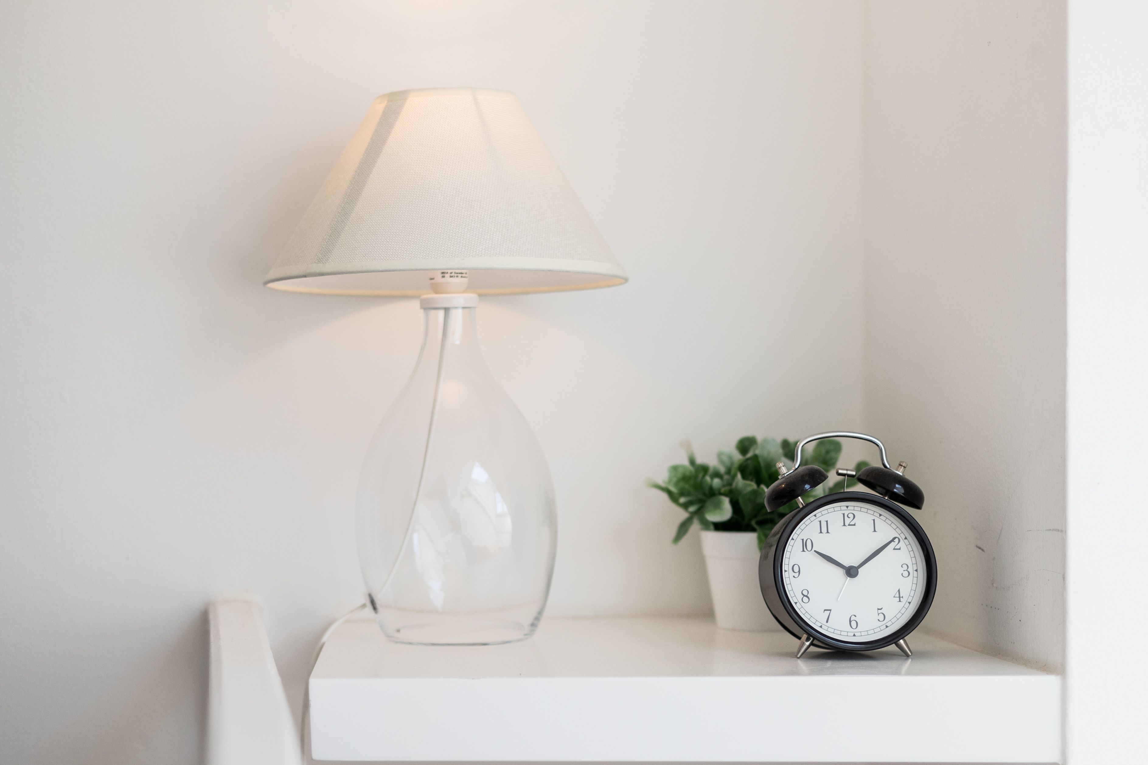 A clear glass lamp with a white shade and a black alarm clock on a white shelf.