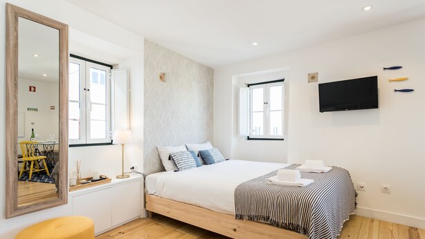 Een moderne slaapkamer met een bed, een grote spiegel, een flatscreen televisie en een eetgedeelte zichtbaar door een open deur.