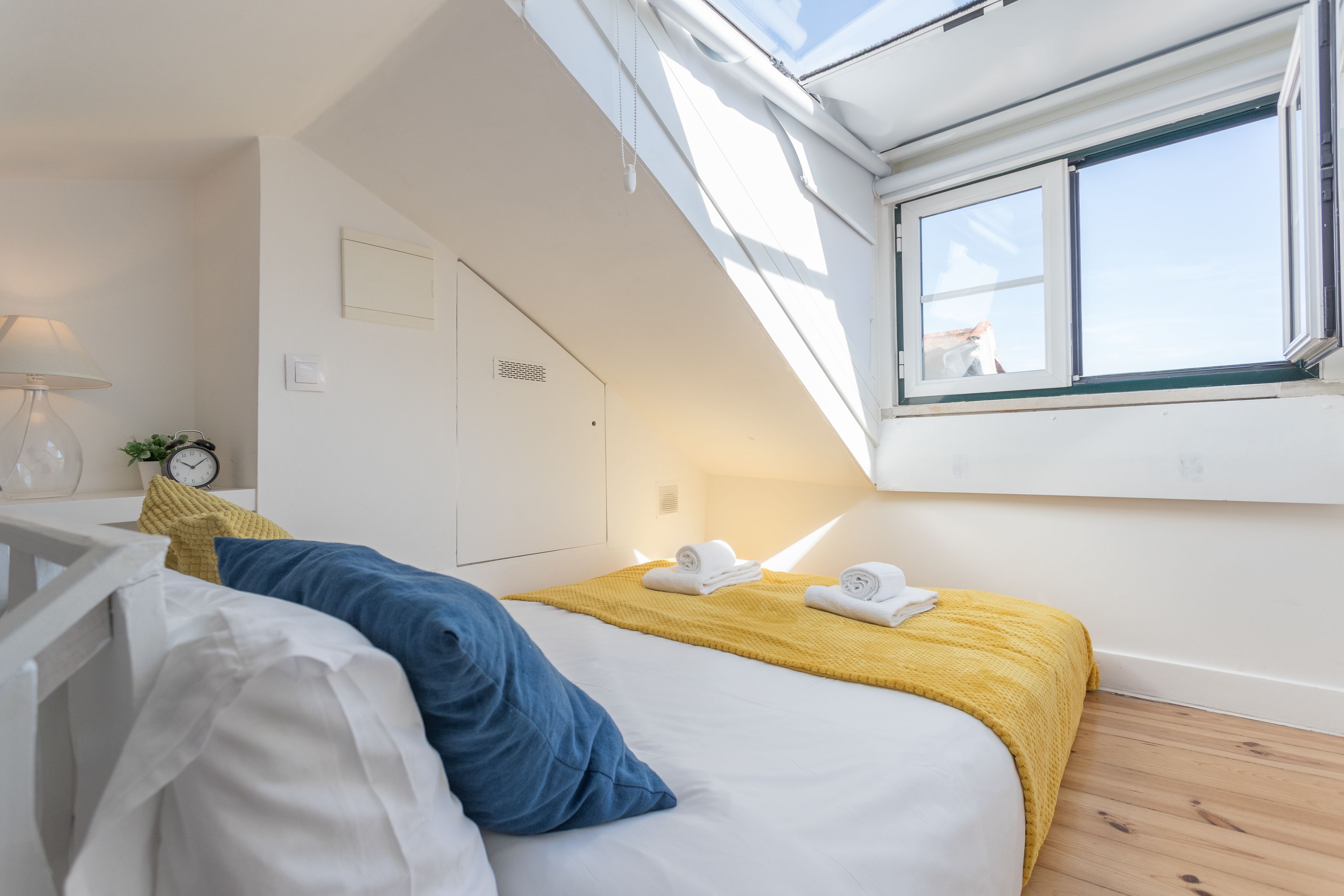 A bedroom with a skylight, a bed with white and blue bedding, a yellow throw blanket, and a bedside table with a lamp and a clock.