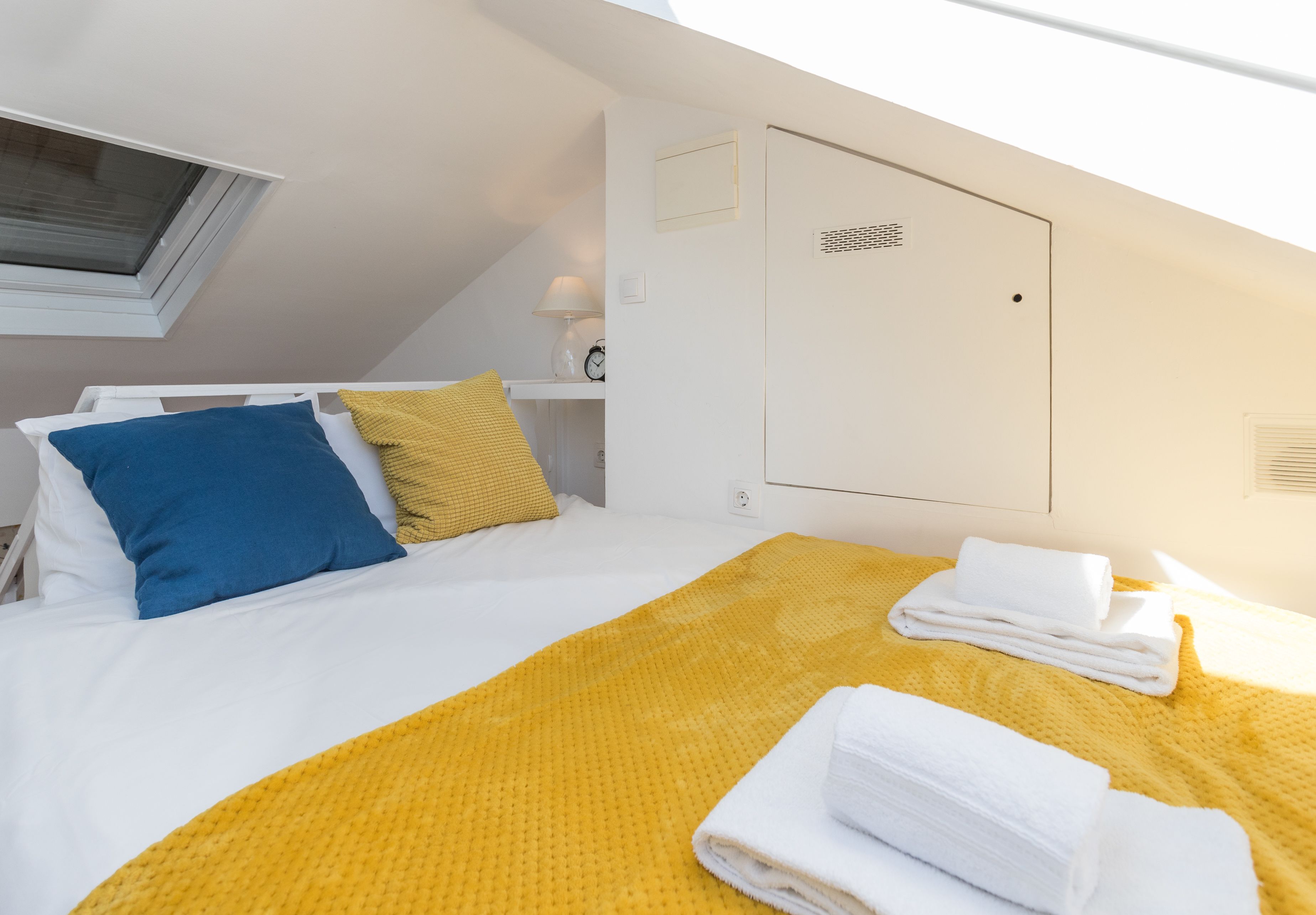 Une chambre d’hôtel avec un lit, une couverture jaune, des serviettes blanches, un oreiller bleu et une fenêtre avec des stores.