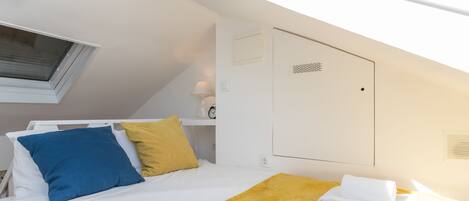 Une chambre d’hôtel avec un lit, une couverture jaune, des serviettes blanches, un oreiller bleu et une fenêtre avec des stores.