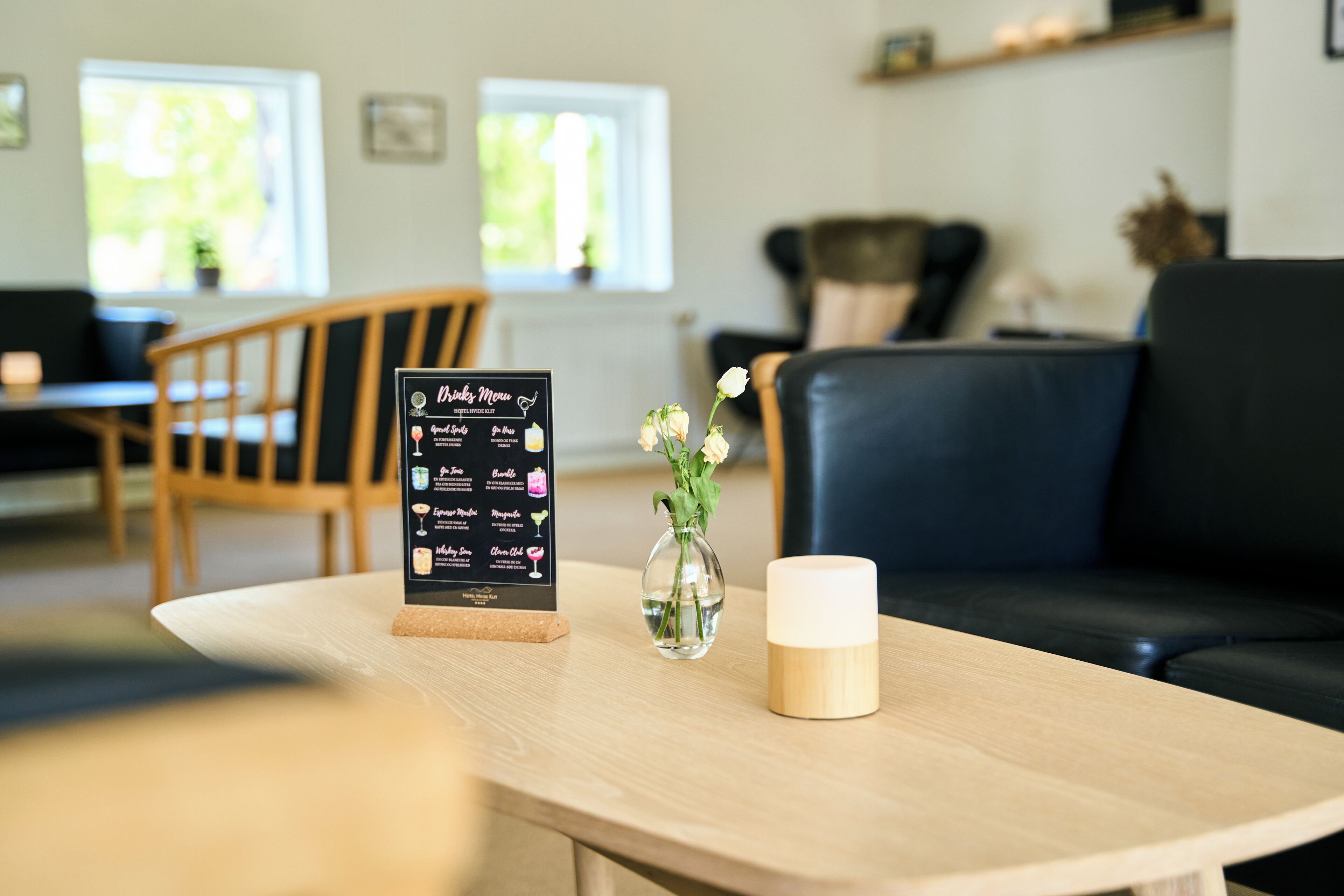 Un tablero de menú con opciones de bebidas, un jarrón con flores y una vela sobre una mesa en una habitación con un sofá y sillones.