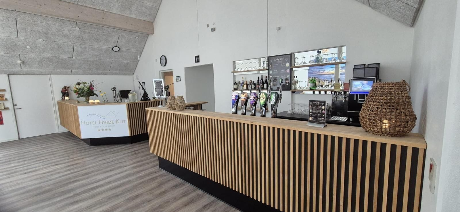 A hotel reception area with a wooden reception desk, a clock on the wall, and a window with a view of the sea.