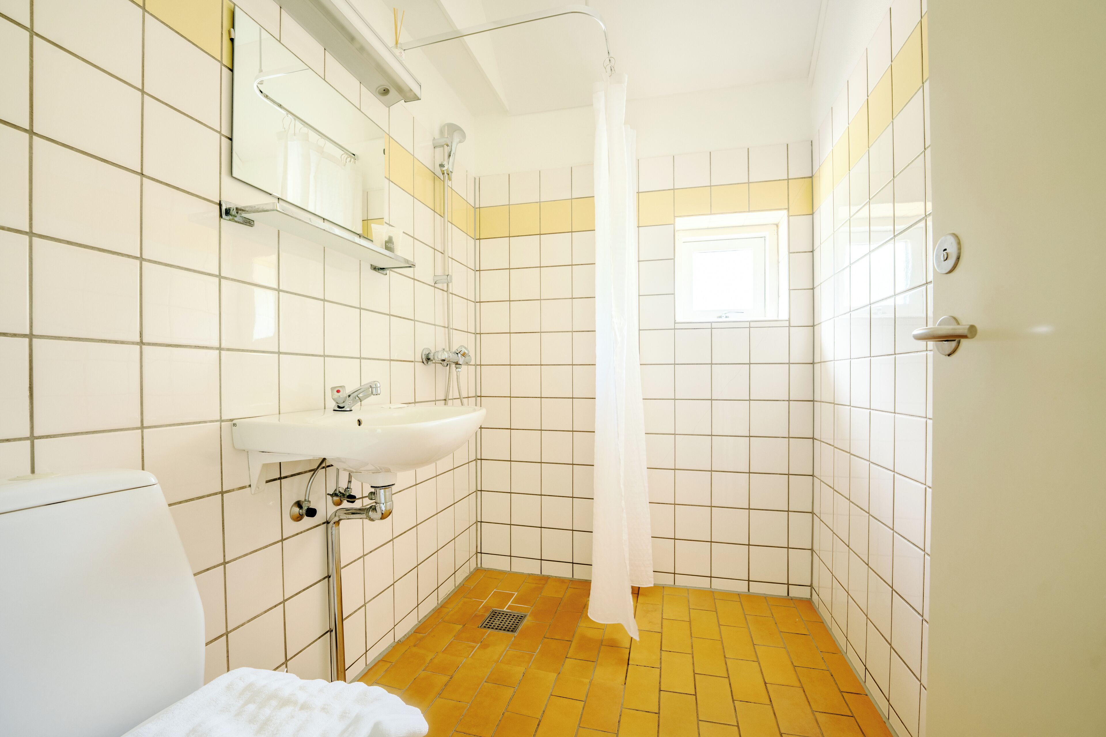 A bathroom with a white sink, mirror, and toilet, a shower with a white curtain, and yellow-tiled walls.