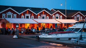 Un restaurante frente al agua con terraza, barcos amarrados cerca y una hilera de edificios rojos con detalles blancos.