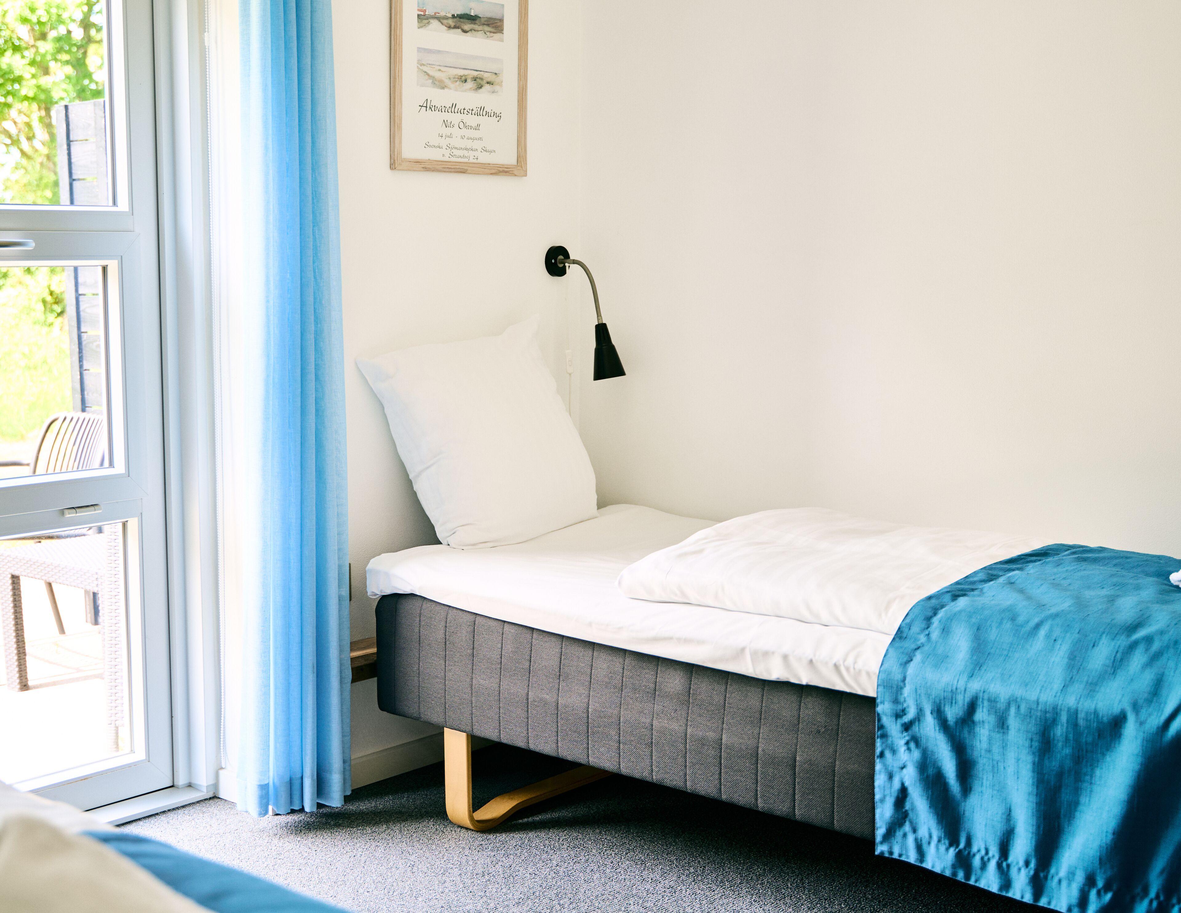 Un dormitorio con una cama, una ventana con cortinas, una lámpara de pared y un cuadro enmarcado.