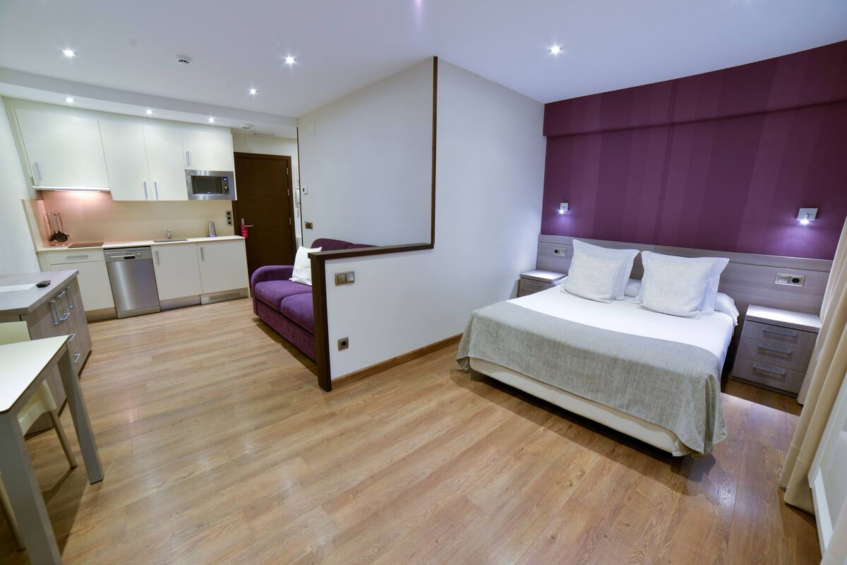 A modern hotel room with a bed, bedside tables, a desk, and a kitchenette.