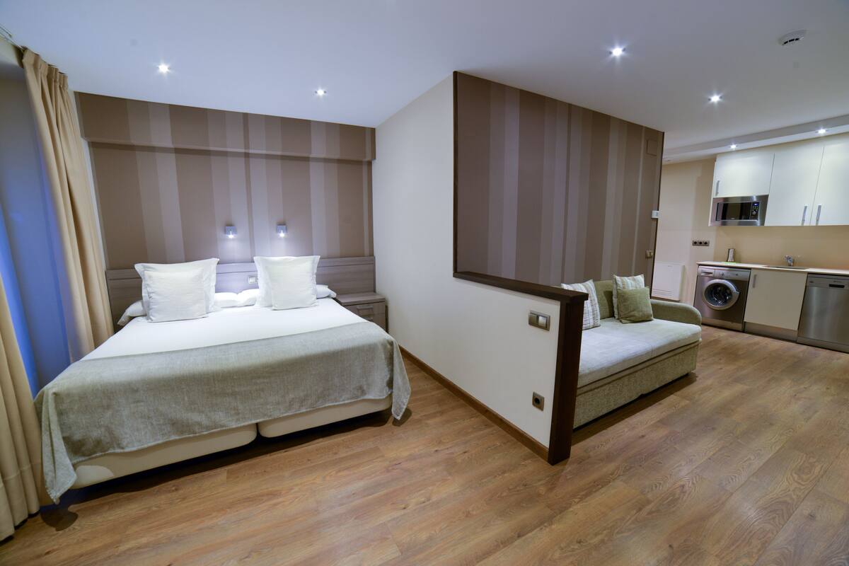 A bedroom with a bed, bedside tables, a sofa, and a kitchenette with a microwave and washing machine.