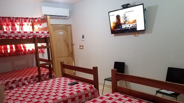 A room with three bunk beds, each with a red and white checkered bedspread. There is a television mounted on the wall displaying a movie poster. - Sharp Guest House (Accra)