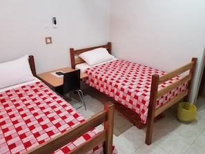 Una habitación pequeña y limpia con dos camas individuales, un escritorio de madera, una silla y un pequeño cubo de basura.
