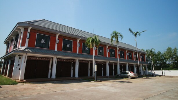 A two-story building with a red facade and a grey roof, featuring multiple garage doors and a parked car. - BarandBed URT (Phunphin)