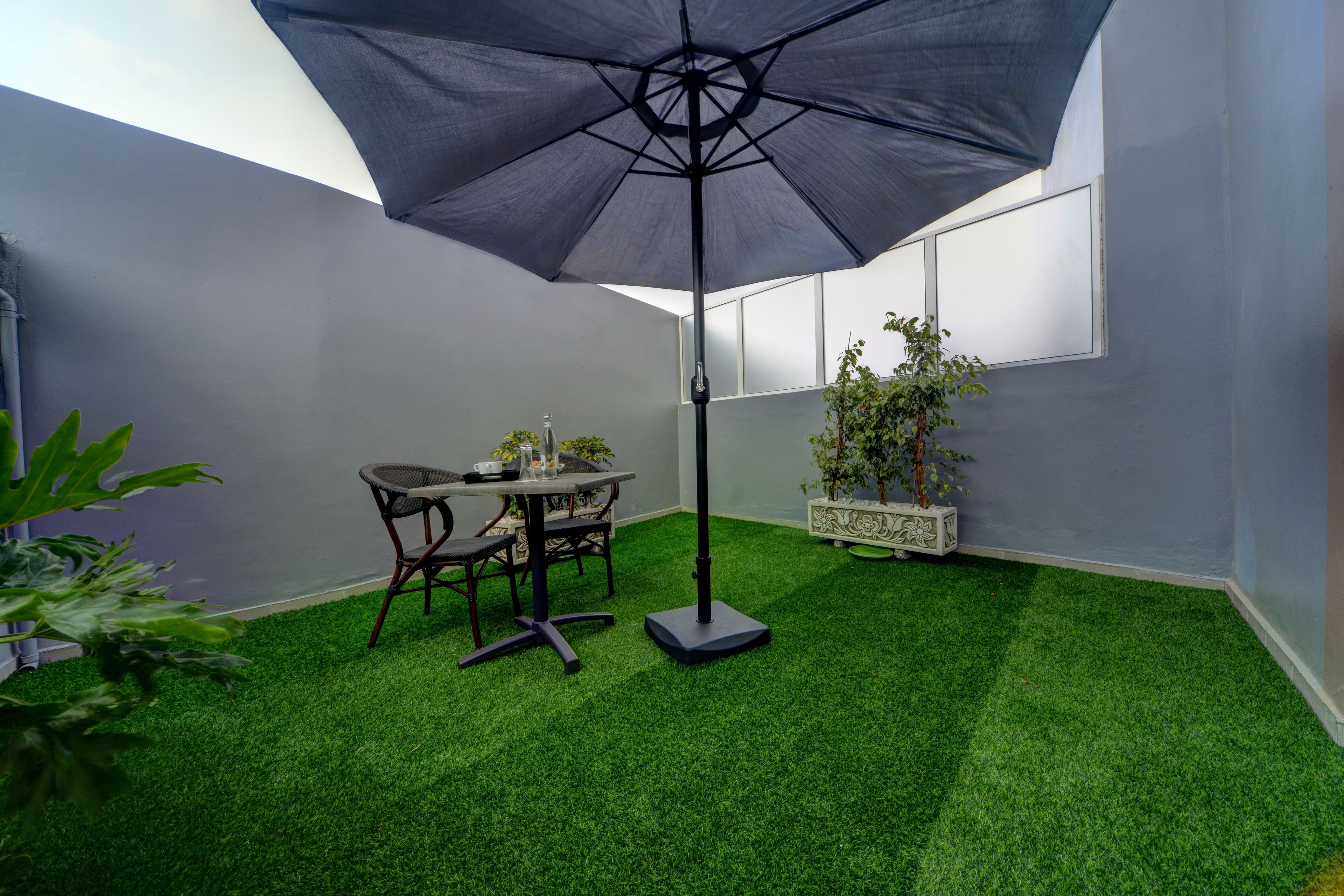 A patio area with artificial grass, a table and chairs, a large umbrella, and potted plants.