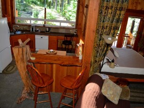 A rustic cabin interior with a wooden dining table, two wooden chairs, a small kitchen area with a sink and a refrigerator, and a large window offering a view of the forest. - Lanzarotta Bed and Breakfast (Eugene)