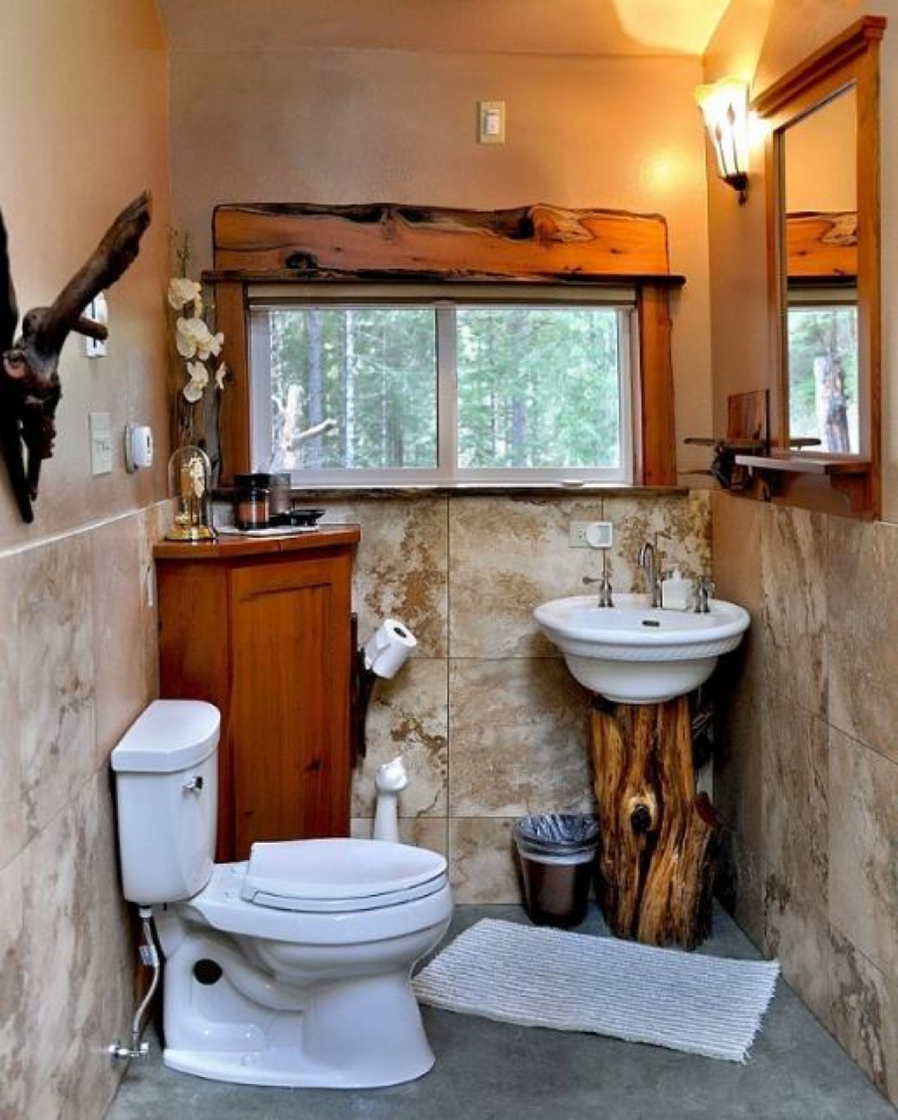 A bathroom with a rustic design, featuring a wooden vanity, a white toilet, a sink with a wooden base, and a window with a view of trees.