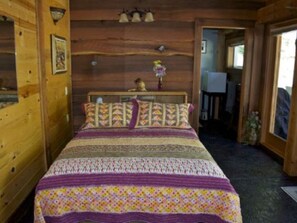 A wooden cabin bedroom with a bed, a wooden headboard, a wall-mounted light fixture, and a door leading to another room. - Lanzarotta Bed and Breakfast (Eugene)