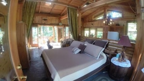 A wooden cabin bedroom with a bed, curtains, a small table with a lamp, and a view of the outdoors. - Lanzarotta Bed and Breakfast (Eugene)
