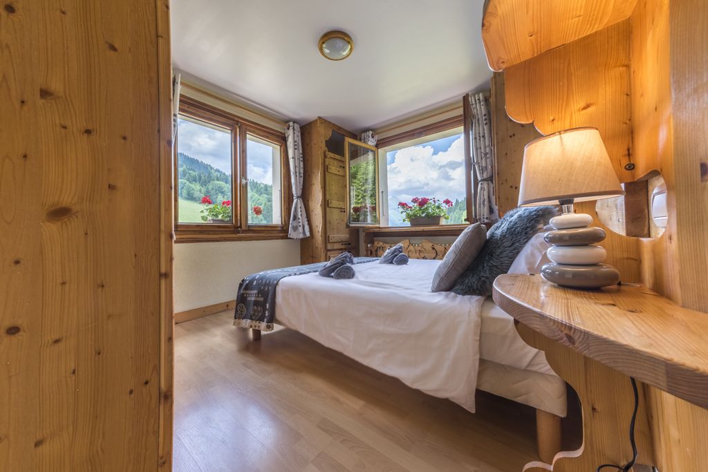 A wooden-framed bedroom with a bed, a bedside table with a lamp, and a view of greenery through large windows.