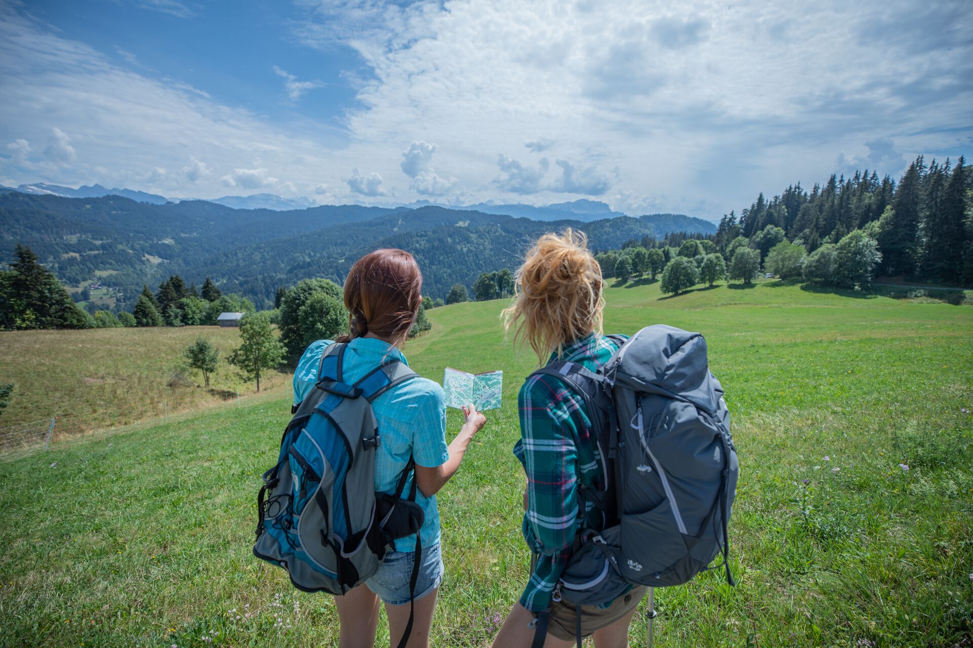Due persone con zaino in spalla osservano una mappa in un prato, con montagne sullo sfondo.