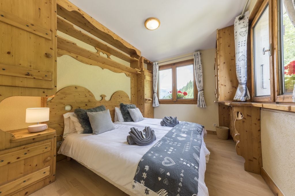 A wooden bedroom with a bed, a nightstand, a window with curtains, and a view of greenery outside.
