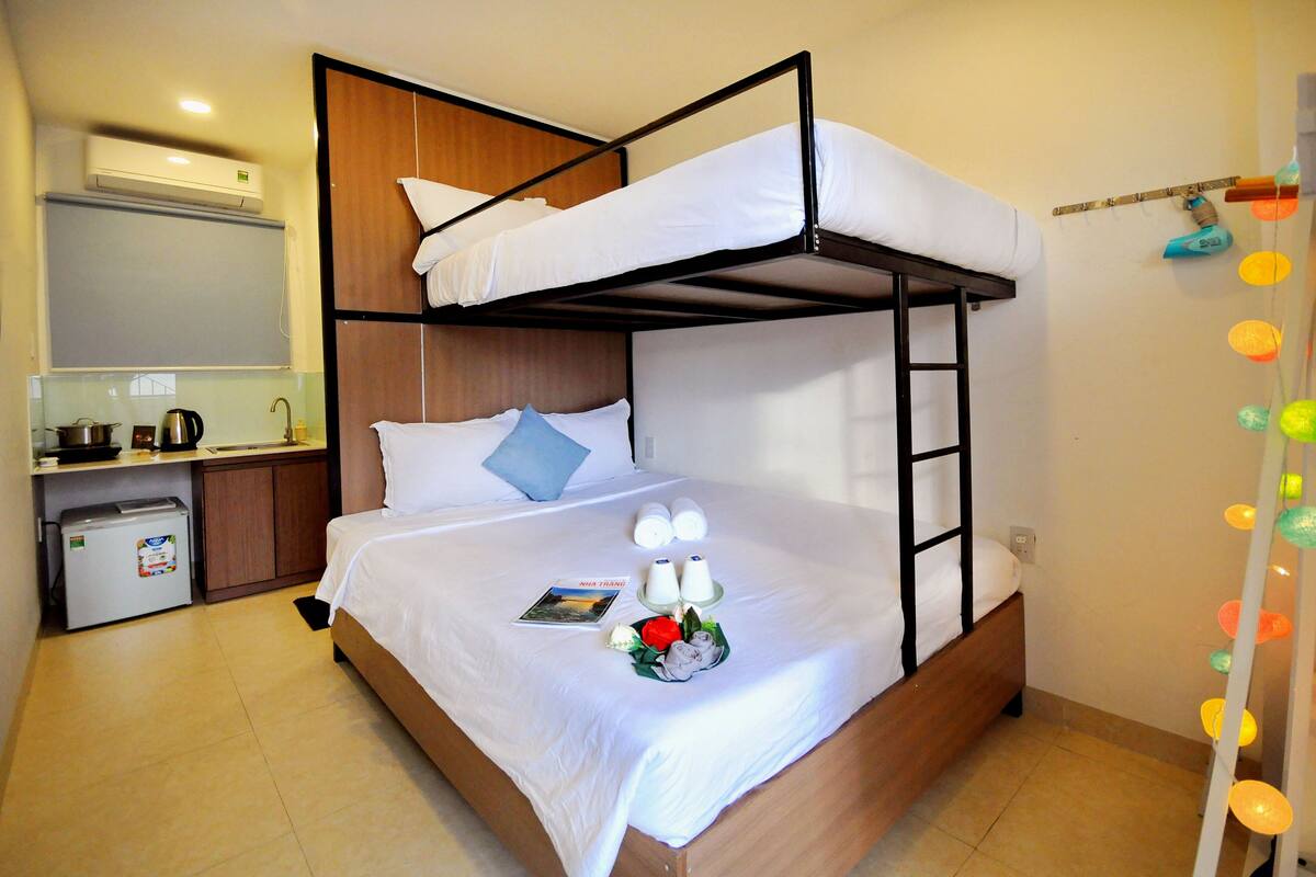 A bunk bed room with a kitchenette, a small fridge, and a bedside table with a lamp.