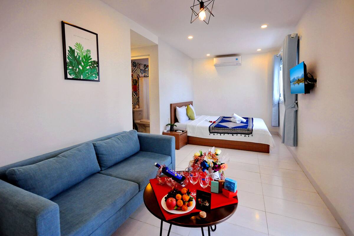 A modern hotel room with a blue sofa, a neatly made bed, a small dining table with fruit and drinks, and a TV mounted on the wall.