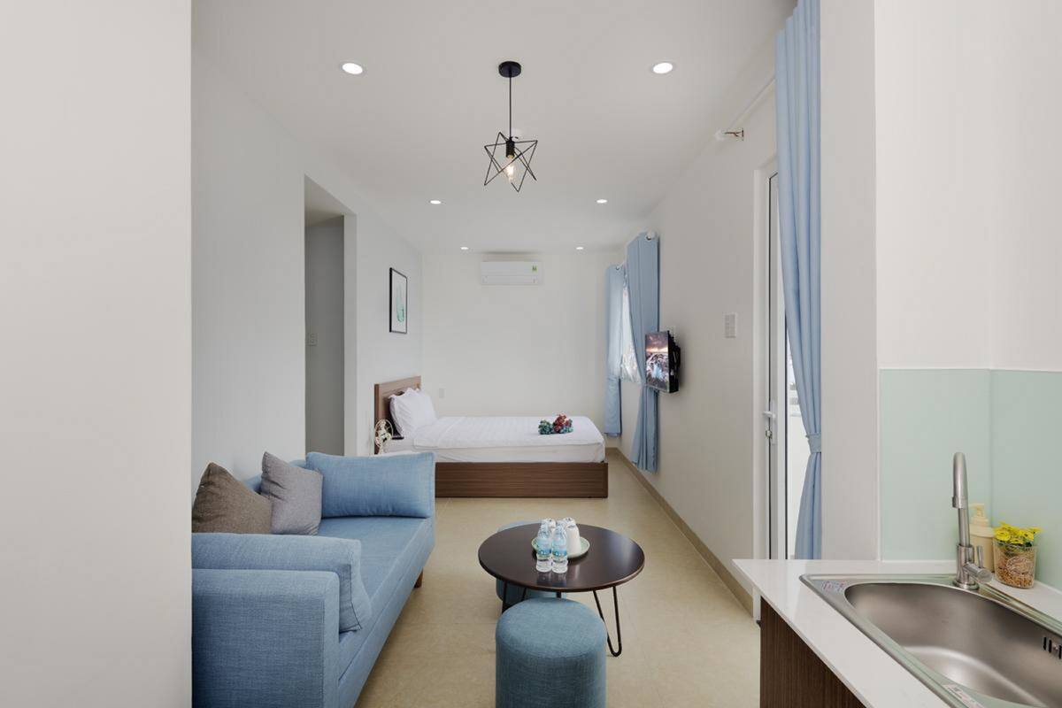 A modern hotel room with a bed, a sofa, a small round table, and a kitchenette area.