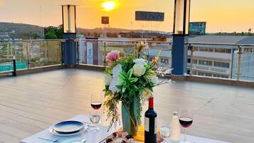 A rooftop dining table set with plates, cutlery, a bottle of wine, and a vase of flowers, overlooking a cityscape at sunset.