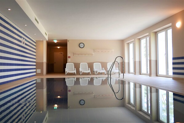 Un área de piscina interior moderna con piso reflectante, asientos junto a la piscina y amplias ventanas que permiten la luz natural.