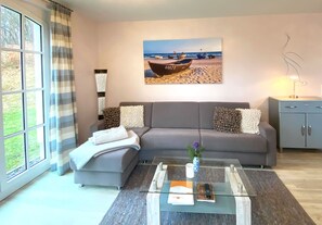 Una sala de estar con un sofá, una mesa de centro de vidrio, un cuadro de barcos en la playa y una lámpara.