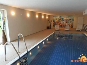 An indoor swimming pool with a blue tiled floor, a metal pool ladder, and white lounge chairs against a wall with a mural.