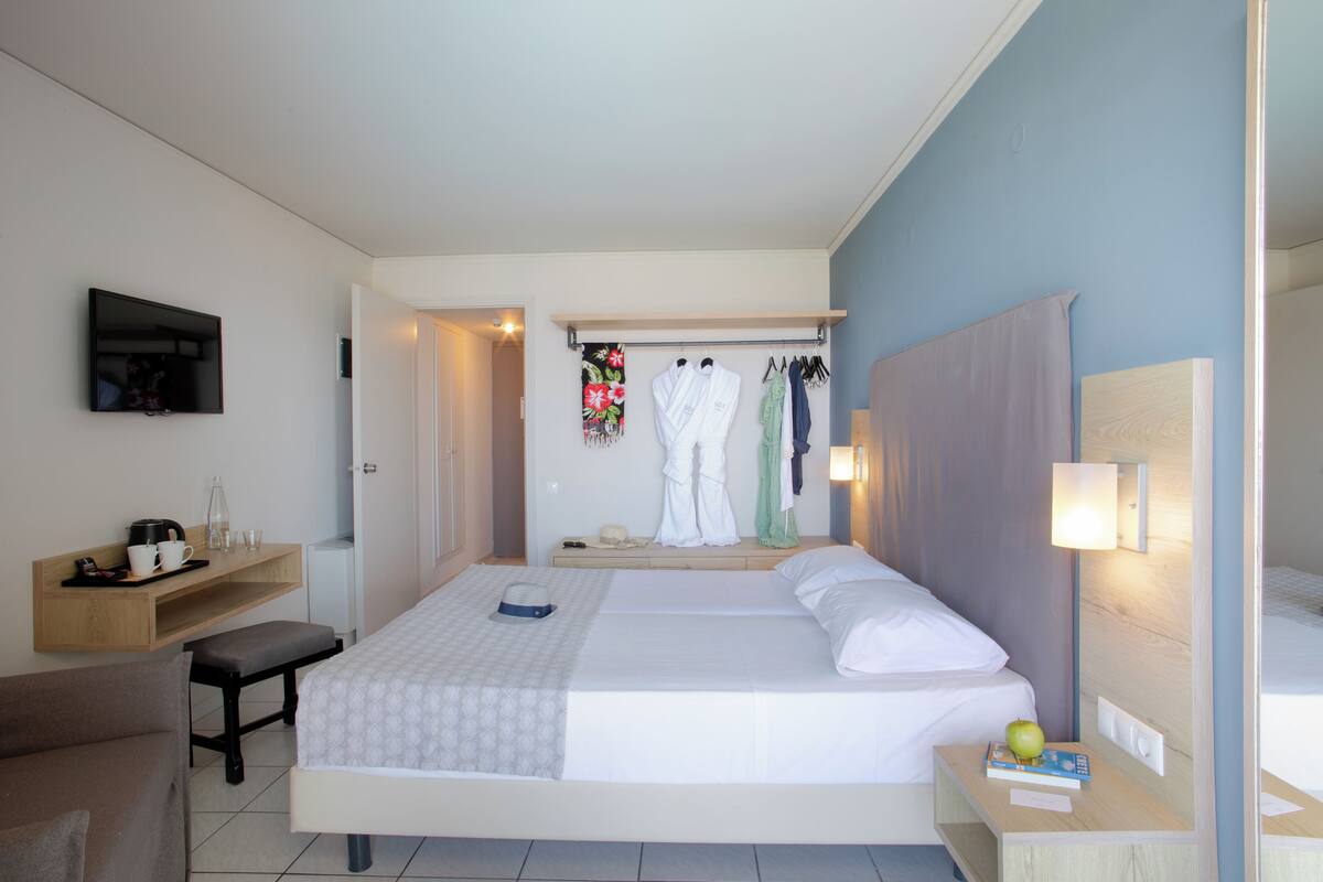 A hotel room with a bed, bedside table, a small desk with a chair, a bench, a TV mounted on the wall, and a clothing rack with robes.