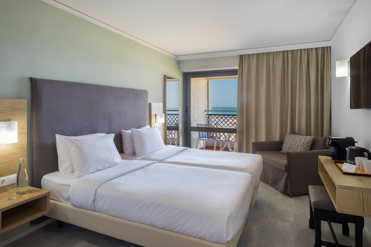 A hotel room with two beds, a desk, a chair, a TV, and a balcony with a view of the sea.