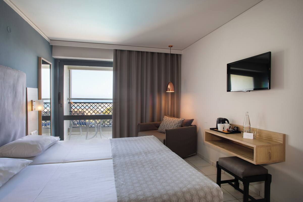 A hotel room with two beds, a sofa, a TV, a small table with a tray, and a balcony with a view.