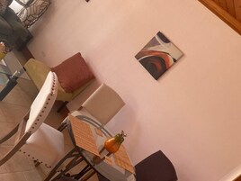 Una habitación con una mesa redonda de vidrio, una silla con asiento de mimbre, un cojín y un cuadro abstracto colgado en la pared.