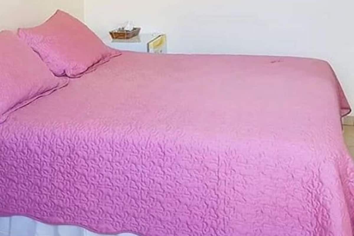 Een bed met een roze deken en kussen, een tafeltje met zakdoos en boek, en een effen muur.