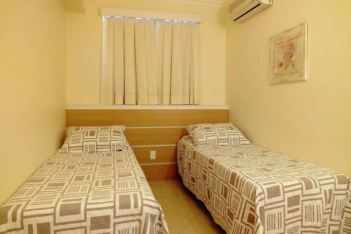 Een kamer met twee eenpersoonsbedden, een raam met zonwering, een airconditioning en een ingelijst schilderij aan de muur.