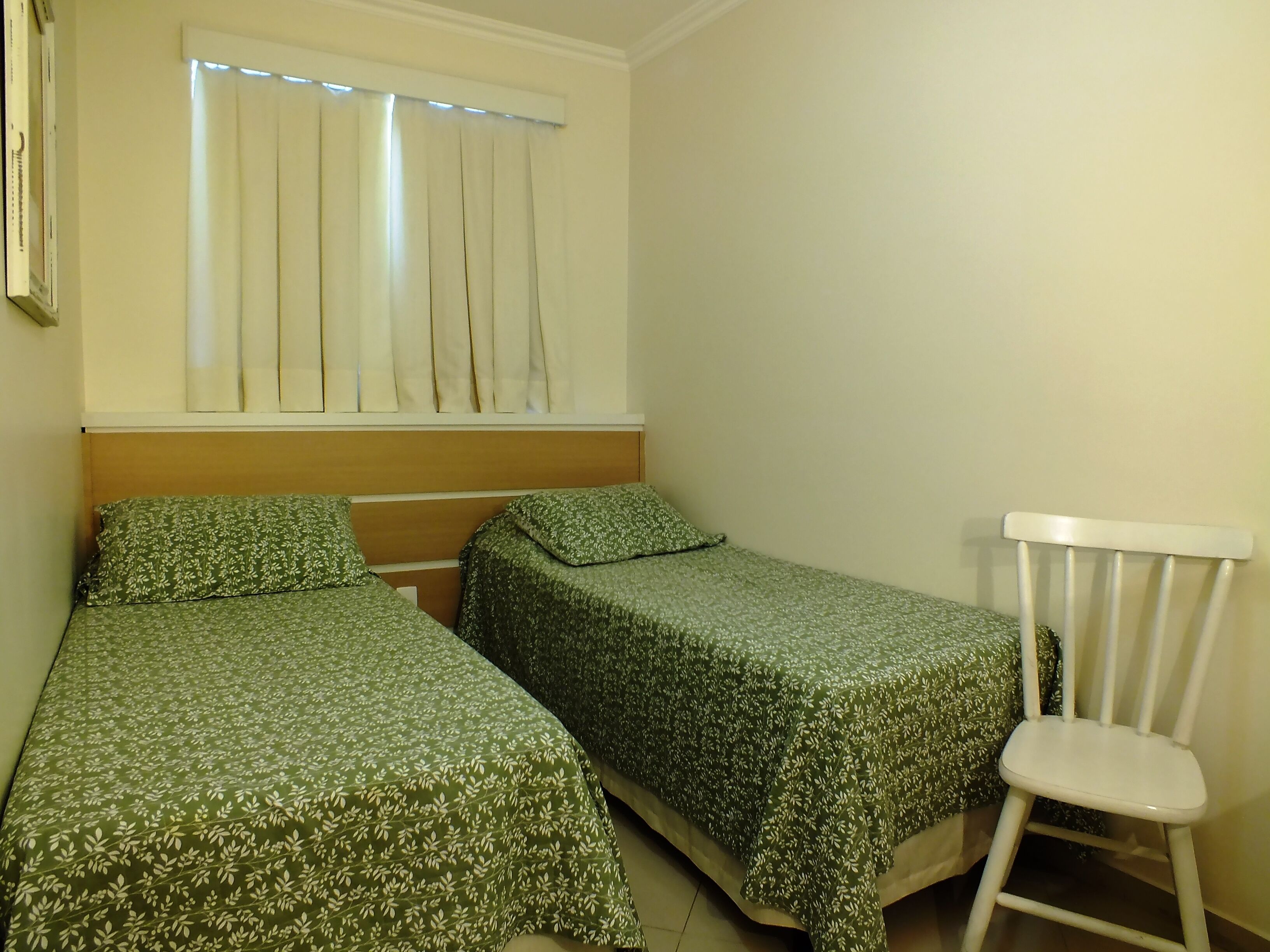 A small, simple room with two single beds, a white chair, and a window with curtains.
