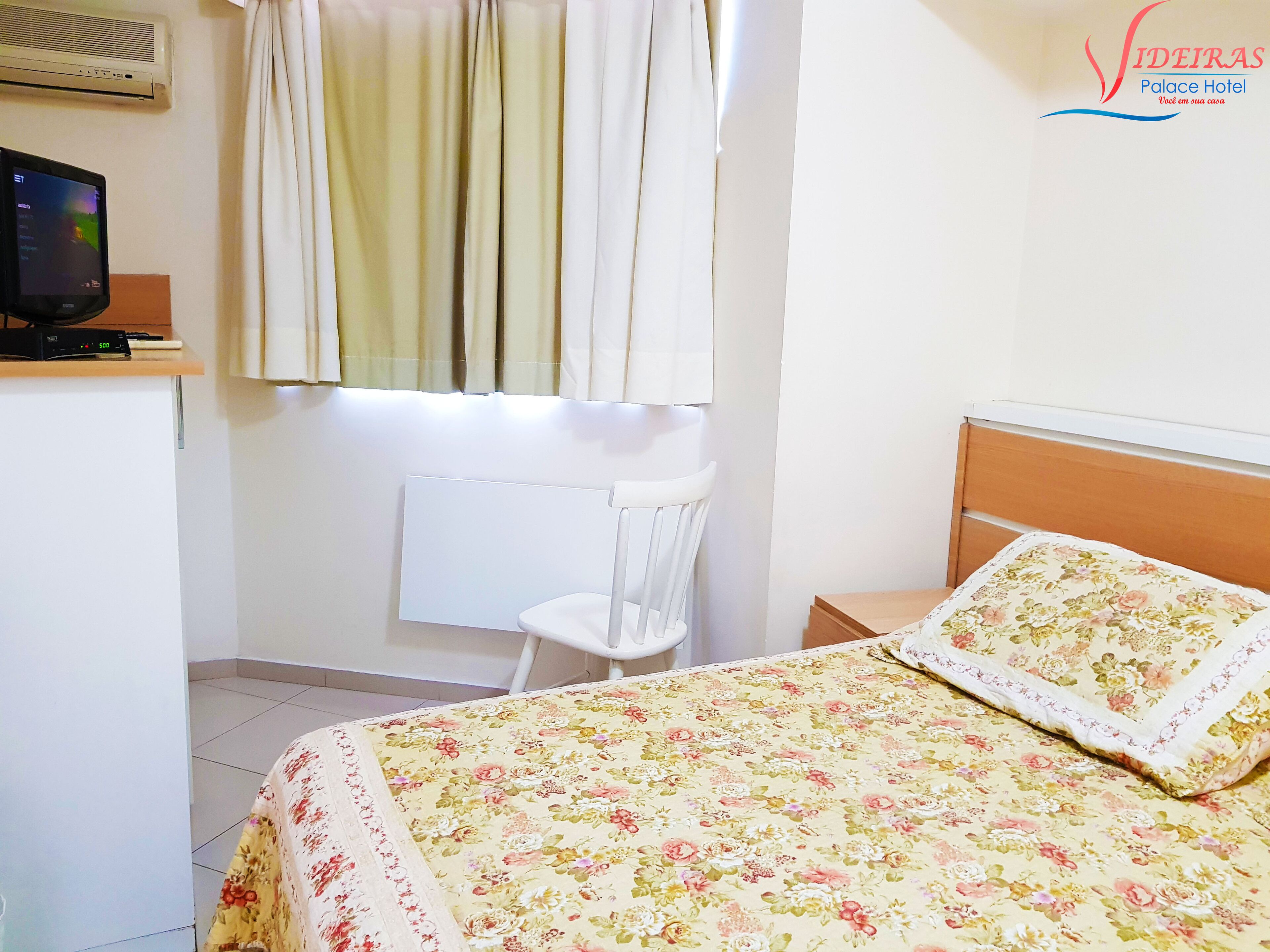 A hotel room with a bed, a small desk with a computer, a radiator, and a window with curtains.